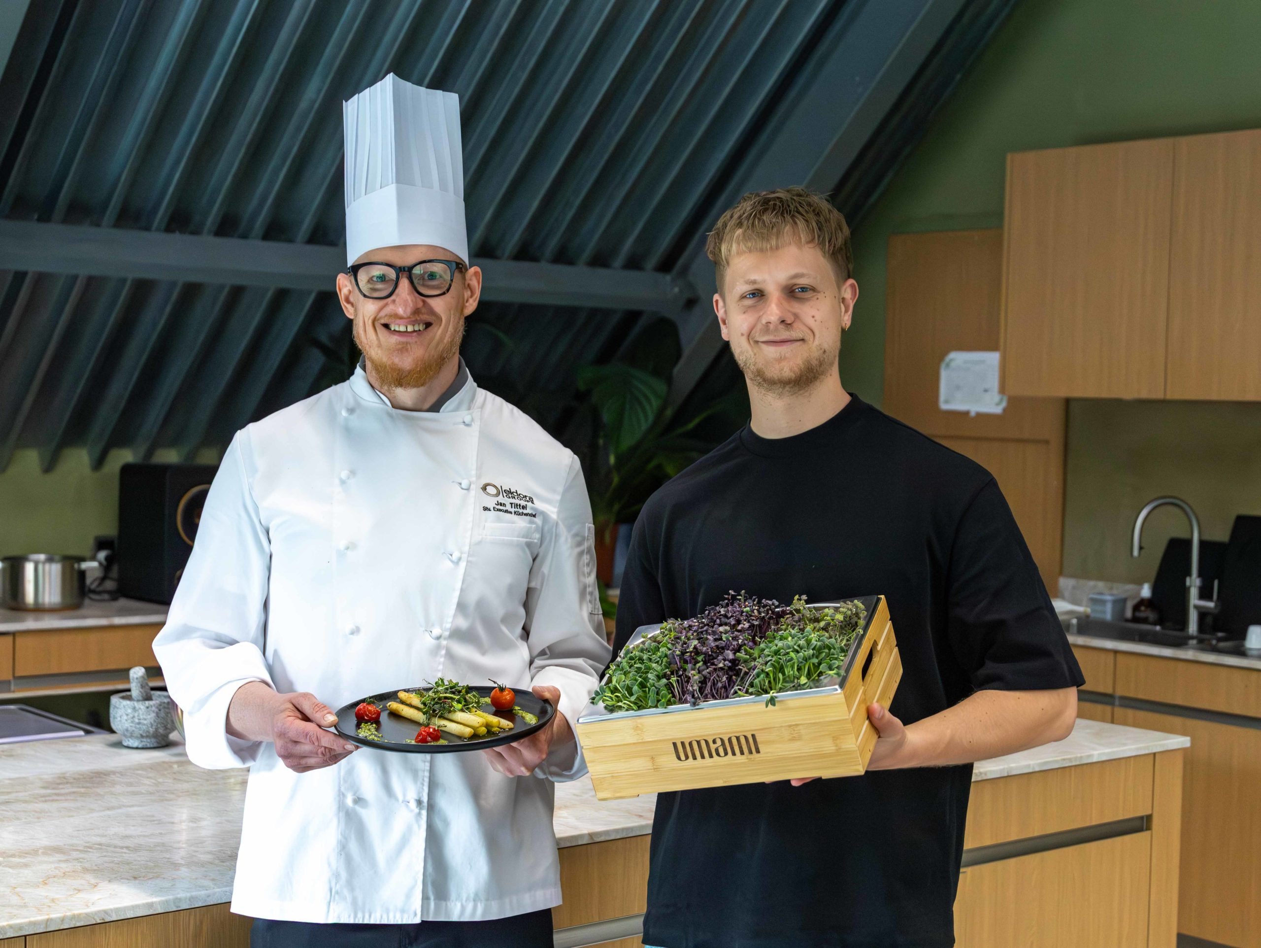 Professioneller Koch in weißer Uniform präsentiert ein Gourmetgericht neben einem Mann mit frischen Microgreens in einer modernen Küche – Fokus auf hochwertige Zutaten, Fine Dining und Umami-Geschmack.