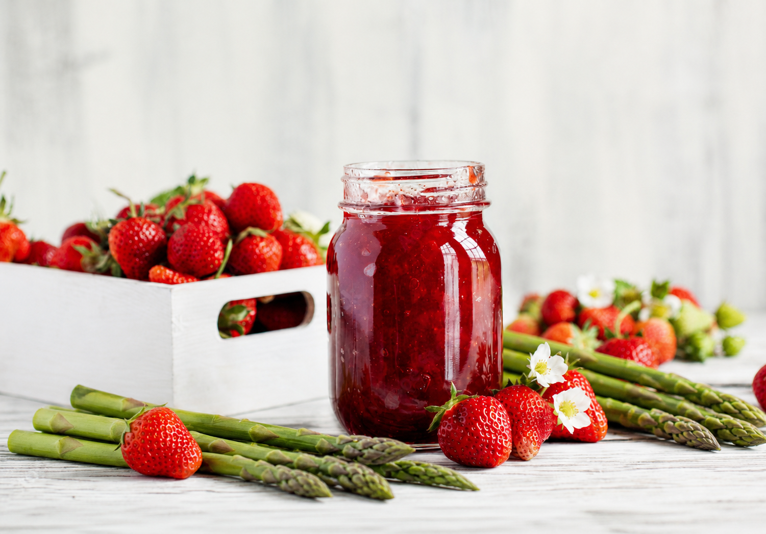 Glas mit hausgemachter Erdbeer-Spargel-Marmelade, umgeben von frischen Erdbeeren, grünem Spargel und weißen Blüten auf hellem Holzuntergrund im natürlichen Licht.