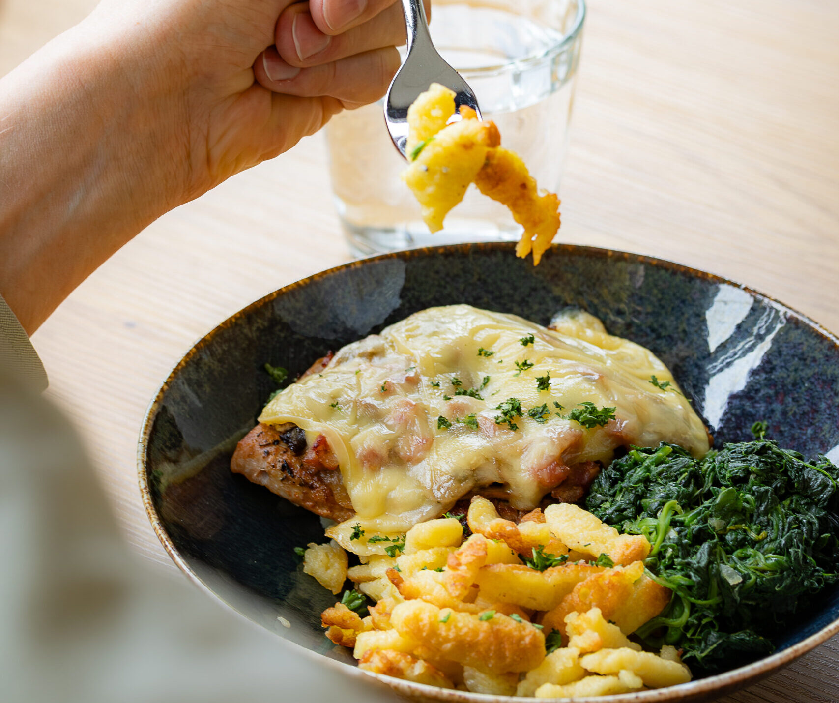 Chäässchnitzel mit Spätzli, Spinat. Fleisch ist von Käsescheibe überbacken und der Teller steht auf einem braunen Tisch